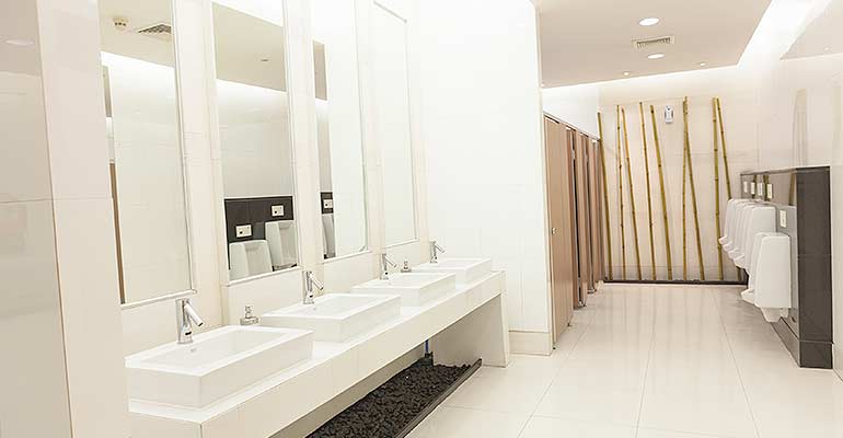 White-tiled public bathroom with row of white modern sinks on left and row of urinals on right.