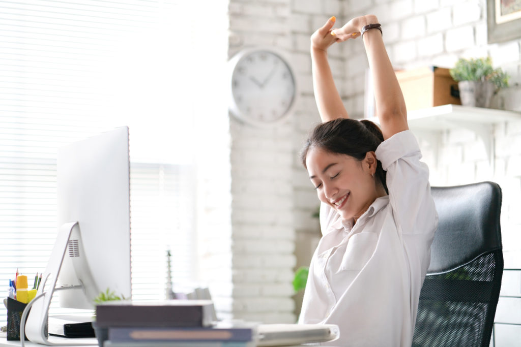Business woman sitting at her desk stretching and smillng; a relaxed, happy look on her face.