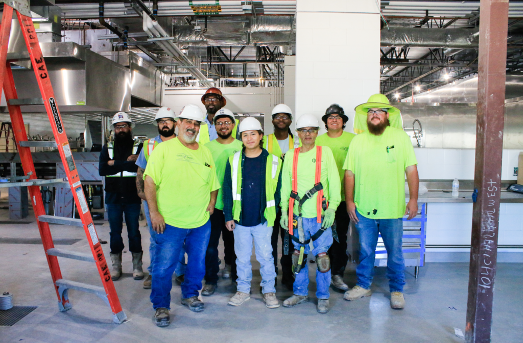 Team of gas line technicians in bright yellow/green t-shirts and white hard hats, standing together in a large industrial warehouse.